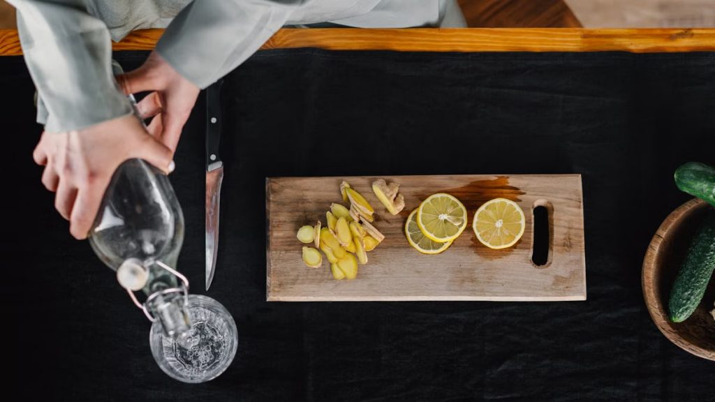 Top view of hands pouring water into a glass with lemon and ginger on a cutting board.