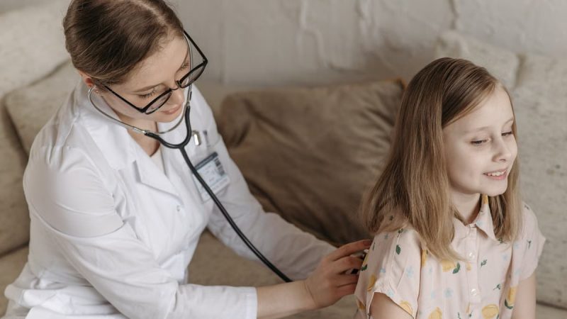A doctor with a stethoscope examining a smiling girl during a medical check-up at home.